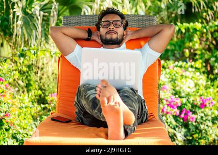 handsome stylish indian man summer vacation in tropical paradise relaxing on an orange deckchair against a background of green tropical trees Stock Photo
