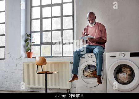 Bald man using tablet PC sitting on washing machine at home Stock Photo