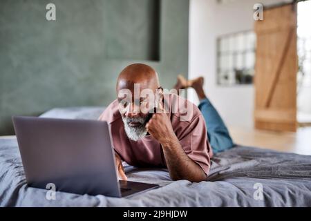 Mature man with white beard using laptop lying on bed at home Stock Photo