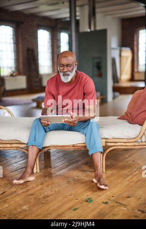 Mature man with white beard using tablet PC sitting on sofa at home Stock Photo