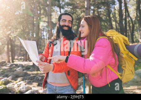 Happy bearded man with map looking at girlfriend wearing backpack in forest Stock Photo
