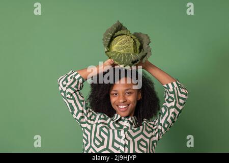 Happy woman with Afro hairstyle holding cabbage on head against green background Stock Photo