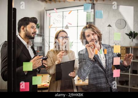 Concentrated male and female entrepreneurs with clipboard and digital tablet writing notes on glass wall at modern office. Group of multiethnic business people having brainstorm session in boardroom. Stock Photo