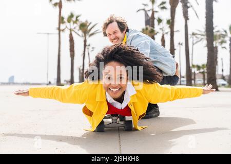 Happy man with playful woman lying on skateboard at footpath Stock Photo