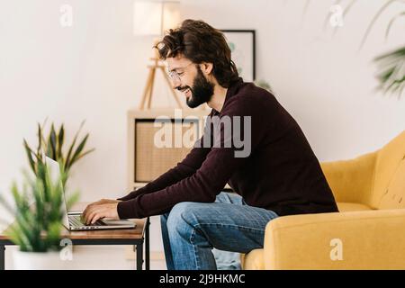 Smiling bearded man using laptop sitting on sofa at home Stock Photo