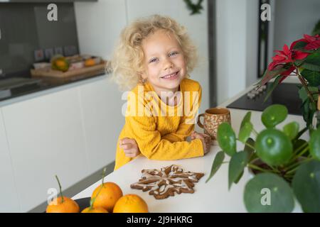 Smiling blond girl with gingerbread leaning on kitchen island Stock Photo