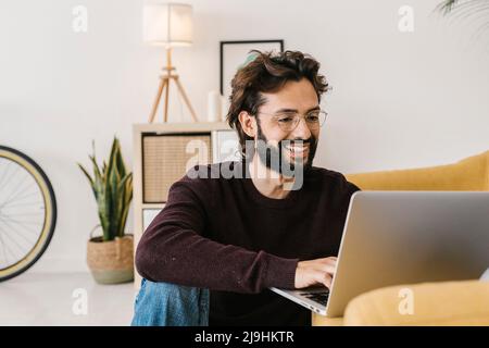 Smiling bearded man using laptop sitting by sofa at home Stock Photo