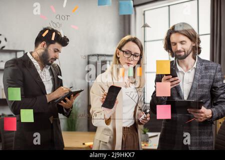 Teamwork of multiethnic managers in formal clothes using modern tablet, smartphone and clipboard during productive meeting in spacious office. Concept of digital devices, people and business. Stock Photo