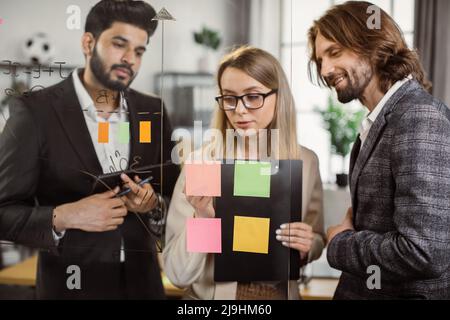 Team of business partners with clipboard and digital tablet in hands discussing new plan at work conference. Multicultural coworkers writing notes on glass wall while brainstorming new startup ideas. Stock Photo