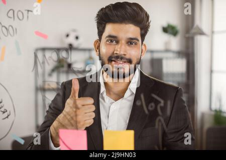 Portrait of executive male manager showing thumb up sign while working on new project at office. Smiling indian man standing in front of glass board and sharing with motivation mood. Stock Photo