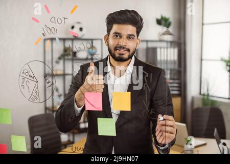 Portrait of executive male manager showing thumb up sign while working on new project at office. Smiling indian man standing in front of glass board and sharing with motivation mood. Stock Photo