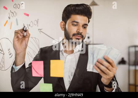 Portrait of male indian employer in formal wear writing on glass board and holding money cash during busy day at work. Young man with calm face counting business profits at modern office. Stock Photo