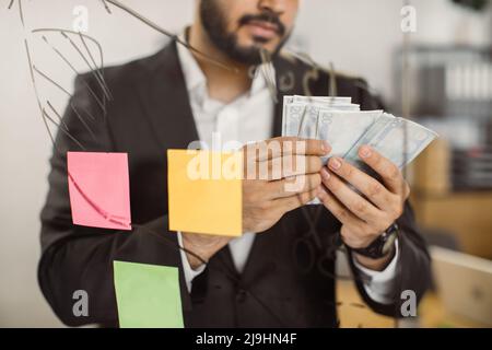 Loose up of concentrated male chief in formal clothes counting money while standing in front of glass board at own company. Salary and business earnings concept. Stock Photo