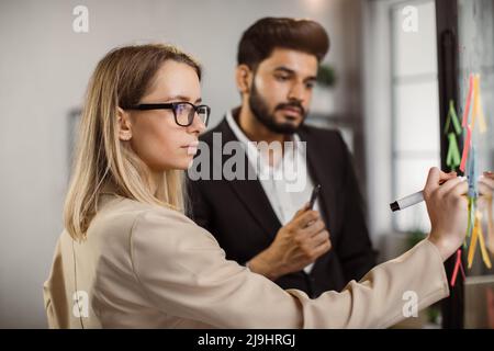 Focused caucasian woman writing marks on glass wall while working together with her indian male colleague in office. Two multiracial managers brainstorming new business ideas during meeting. Stock Photo