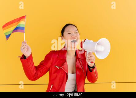 Happy woman holding rainbow flag shouting through megaphone in front of yellow wall Stock Photo