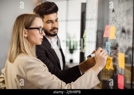 Multicultural group of marketers developing business concept for attracting new customers in company. Young caucasian woman and indian man recording step-by-step actions on glass board. Stock Photo