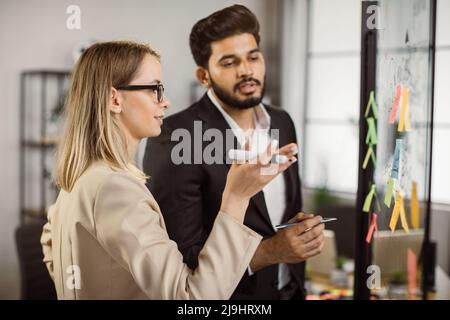 Side view of multi ethnic office managers discussing written information while standing near transparent glass board. Professional company workers considering global business strategy. Stock Photo