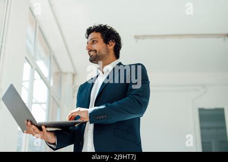 Smiling businessman holding laptop standing in office Stock Photo