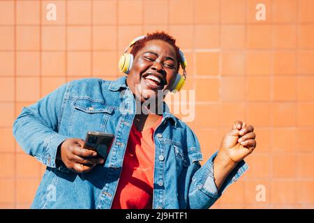 Cheerful woman holding smart phone listening music through wireless headphones enjoying in front of wall Stock Photo