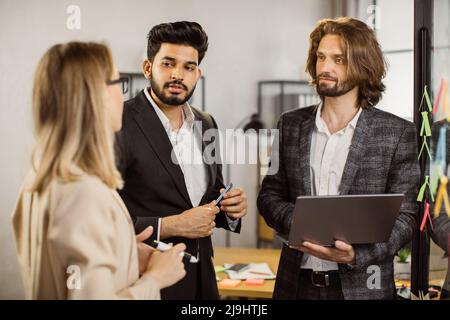 Confident caucasian boss using drawings and reminders on sticky notes while standing near glass board with laptop in hands. Male entrepreneur presenting new ideas to his multiracial employees. Stock Photo