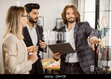 Confident caucasian boss using drawings and reminders on sticky notes while standing near glass board with laptop in hands. Male entrepreneur presenting new ideas to his multiracial employees. Stock Photo