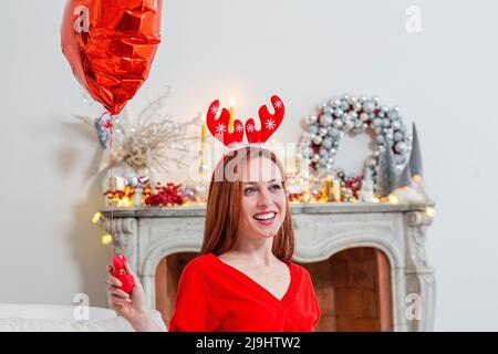 Smiling woman wearing Christmas headdress sitting with red heart shape balloon at home Stock Photo