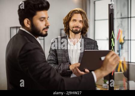 Positive caucasian man in suit holding laptop and looking at camera while his indian business partner writing on glass board. Multi ethnic workmates planning on new strategies in office. Stock Photo