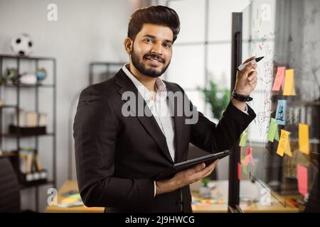 Smiling handsome businessman drawing graphs on glass presentation board in modern office. Male indian manager holding digital tablet in hand and looking at camera. Stock Photo