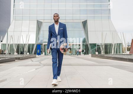 Young businessman holding skateboard and laptop walking in front of office building Stock Photo