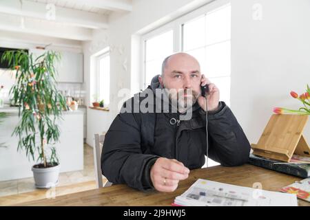 Mature man sitting at home wearing winter jacket making a phone call Stock Photo