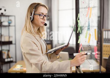 Female office manager in headset making notes on glass board during online conversation with investors at conference hall. Side view of caucasian woman holding portable laptop in hands. Stock Photo