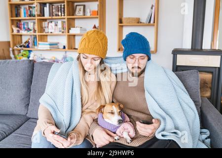 Couple with dog sitting on couch wrapped in blanket looking at smartphone Stock Photo