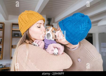 Couple wearing wooly hat at home kissng dog with scarf Stock Photo