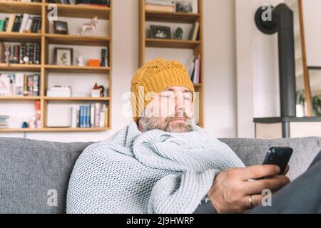 Man wrapped in blanket wearing wooly hat sitting on couch checking smartphone Stock Photo