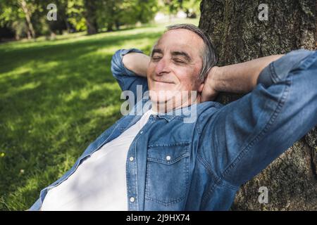 Smiling senior man with eyes closed leaning on tree trunk at park Stock Photo