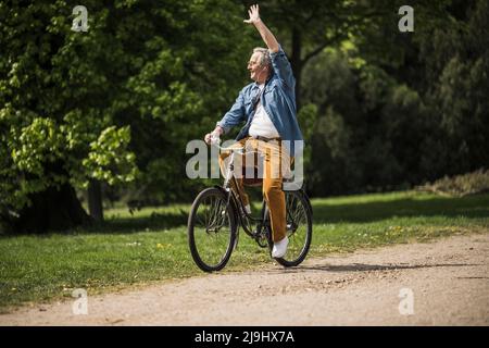 Smiling senior man riding bicycle waving hand at park on sunny day Stock Photo