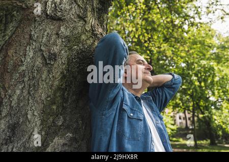 Smiling senior man with hands behind head leaning on tree trunk at park Stock Photo