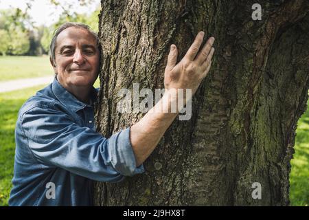 Smiling senior man hugging tree trunk at park Stock Photo