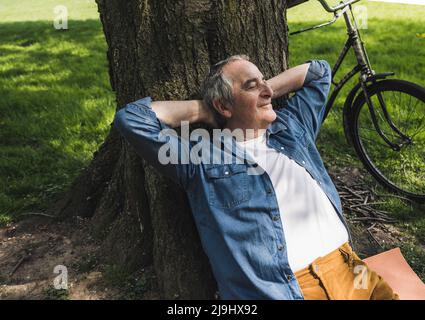 Smiling senior man leaning on tree trunk by bicycle at park Stock Photo