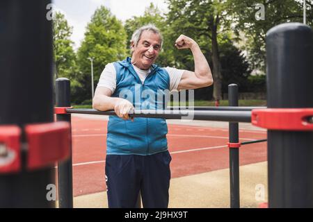 Happy senior man flexing muscles standing by gymnastics bar Stock Photo