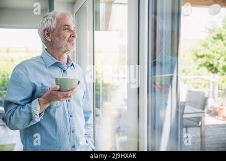 Happy senior man holding coffee cup looking out through window Stock Photo