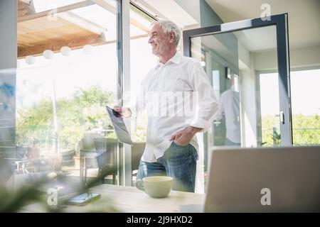 Smiling senior man with tablet PC standing glass window at home Stock Photo