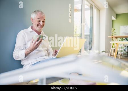 Happy senior man holding coffee cup using laptop at home Stock Photo
