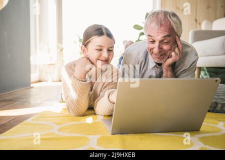Smiling senior man with granddaughter using laptop lying on carpet at home Stock Photo