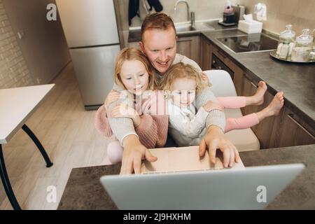 Smiling businessman using laptop sitting with daughters in kitchen at home Stock Photo