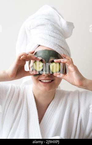 Smiling young woman with face mask covering eyes with cucumber slices in front of wall Stock Photo