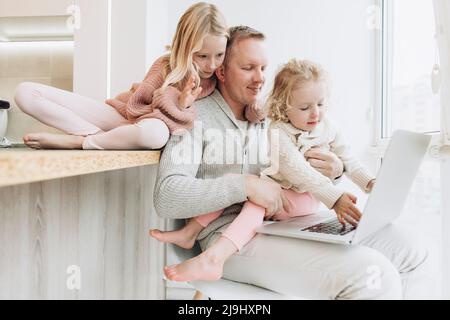 Daughters with father working on laptop sitting by window at home Stock Photo