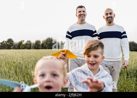 Gay male couple having fun with sons children outdoor - LGBTQ family playing with airplane paper toy - Focus on fathers face Stock Photo