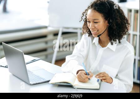 Friendly positive African American woman with curly hair, online sales manager, support service worker, sitting at a work desk in front of a laptop in modern office, making work notes Stock Photo