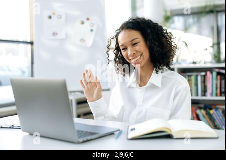 Friendly positive African American girl, ceo, sales manager, sits at a desktop in front of a computer screen with a notepad, holds an video business meeting with a colleague or client, waves, smiles Stock Photo